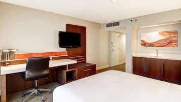 A hotel room with a bed, desk, chair, TV, and artwork on the wall.