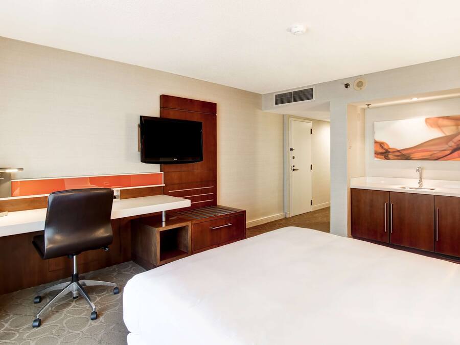 A hotel room with a bed, desk, chair, TV, and artwork on the wall.