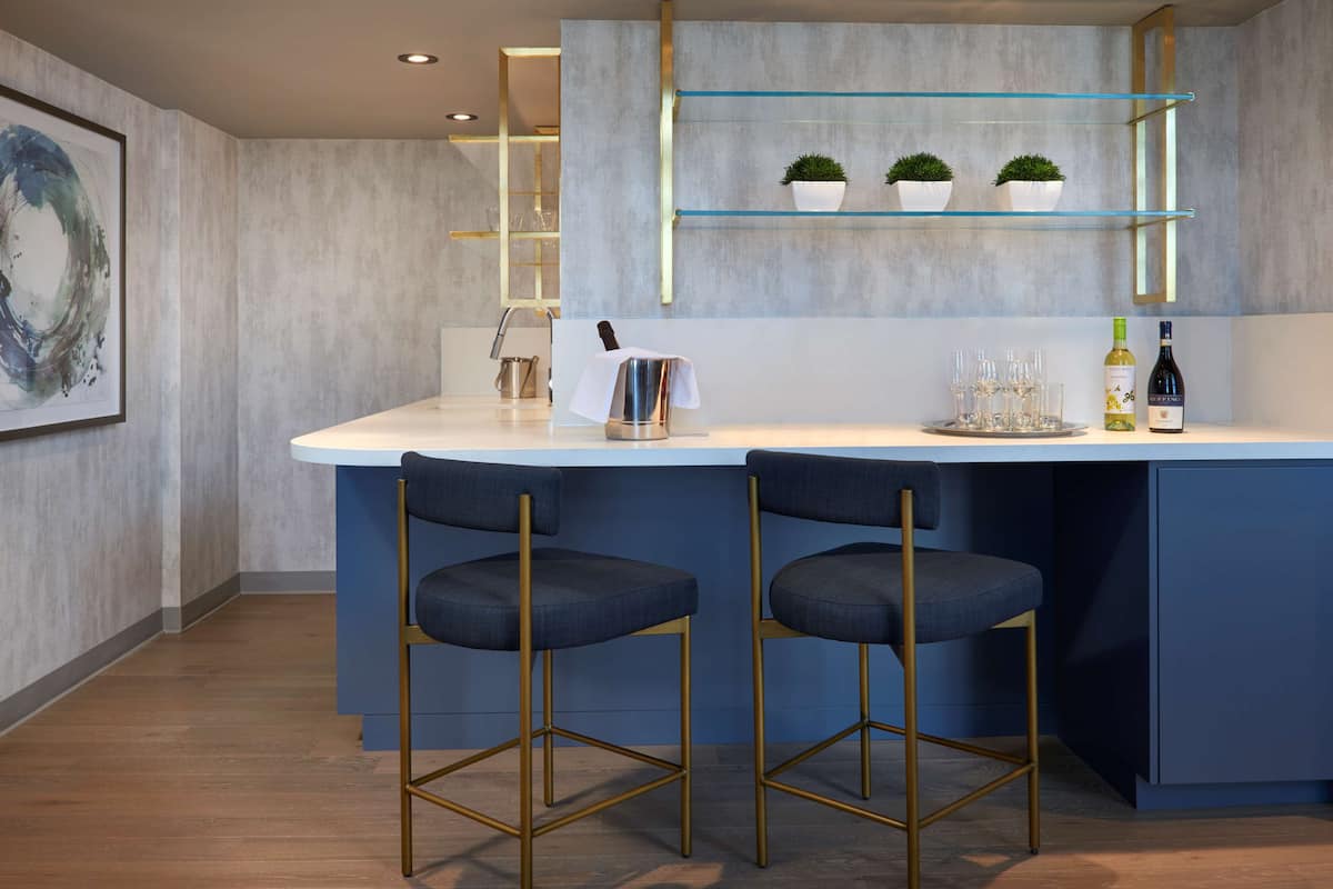 A modern bar area with two bar stools, a white countertop, and glass shelves with potted plants.