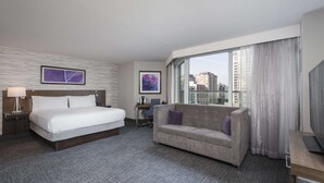Une chambre d’hôtel équipée d’un lit, d’un canapé, d’un bureau et d’une grande fenêtre donnant sur la ville.