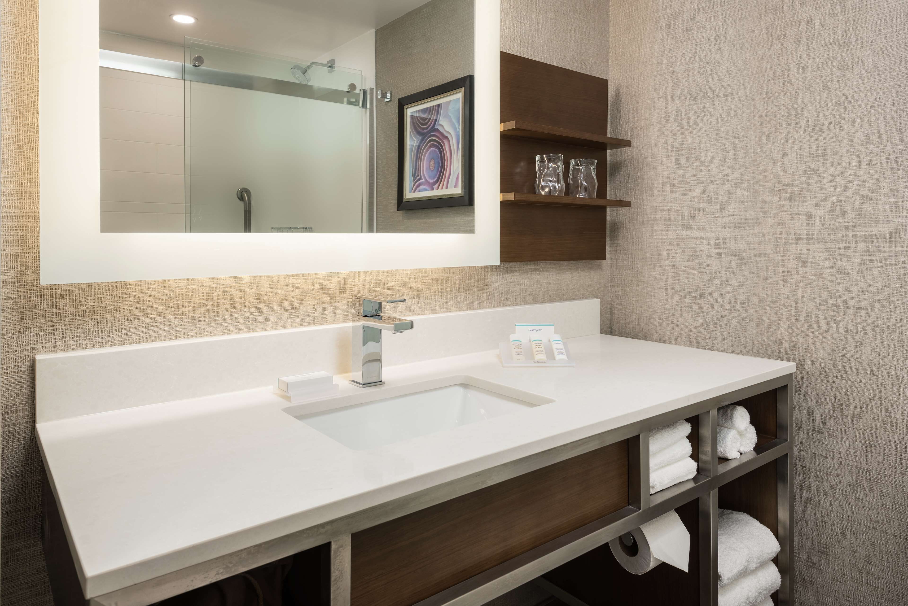 A modern bathroom with a large mirror, a white sink, and a wooden vanity with shelves holding towels and toiletries.