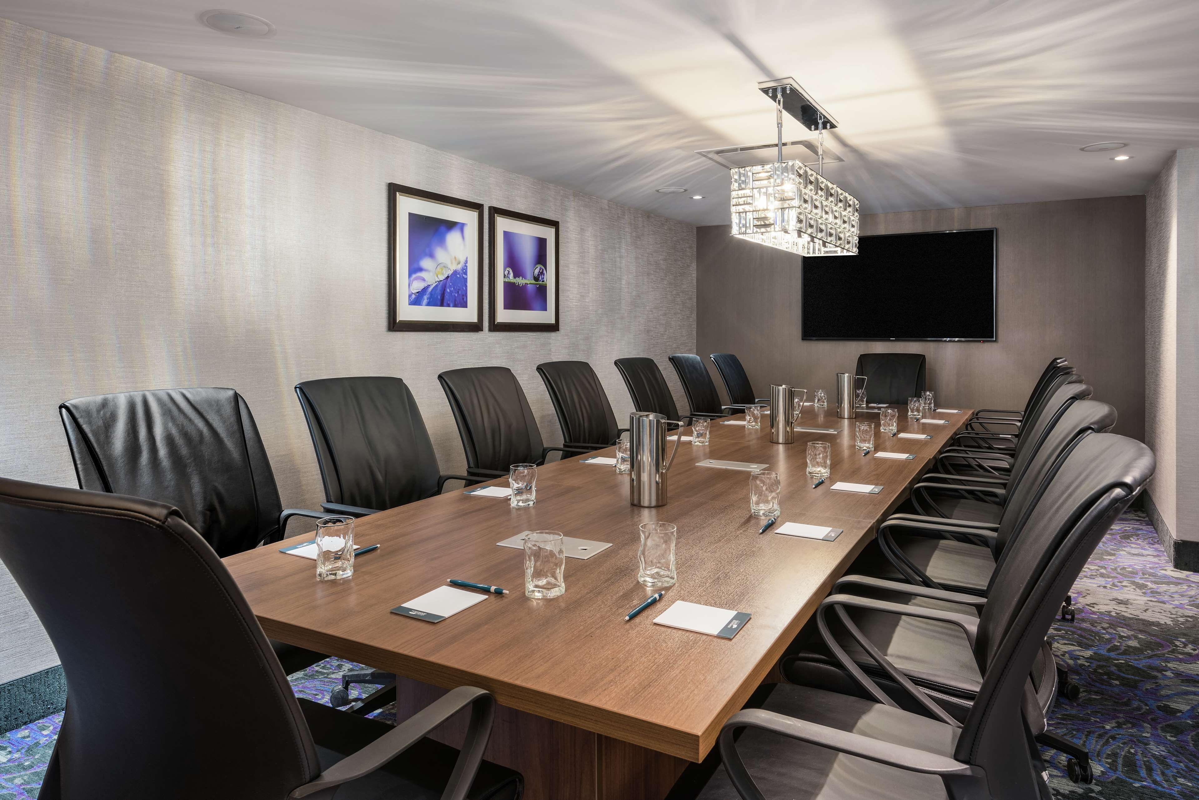 A conference room with a long wooden table, black leather chairs, and a large flat-screen TV on the wall.