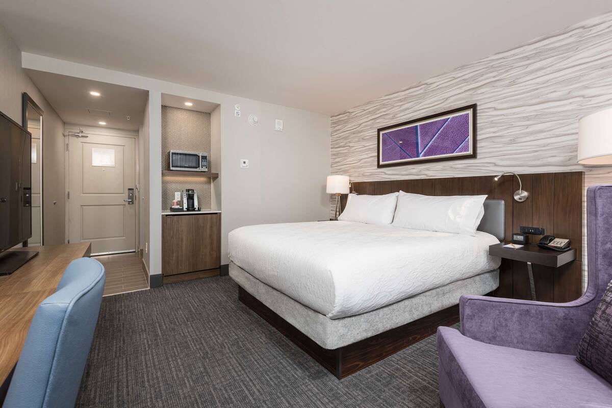 A hotel room with a bed, a desk, a chair, a microwave, a coffee machine, and a wall-mounted TV.