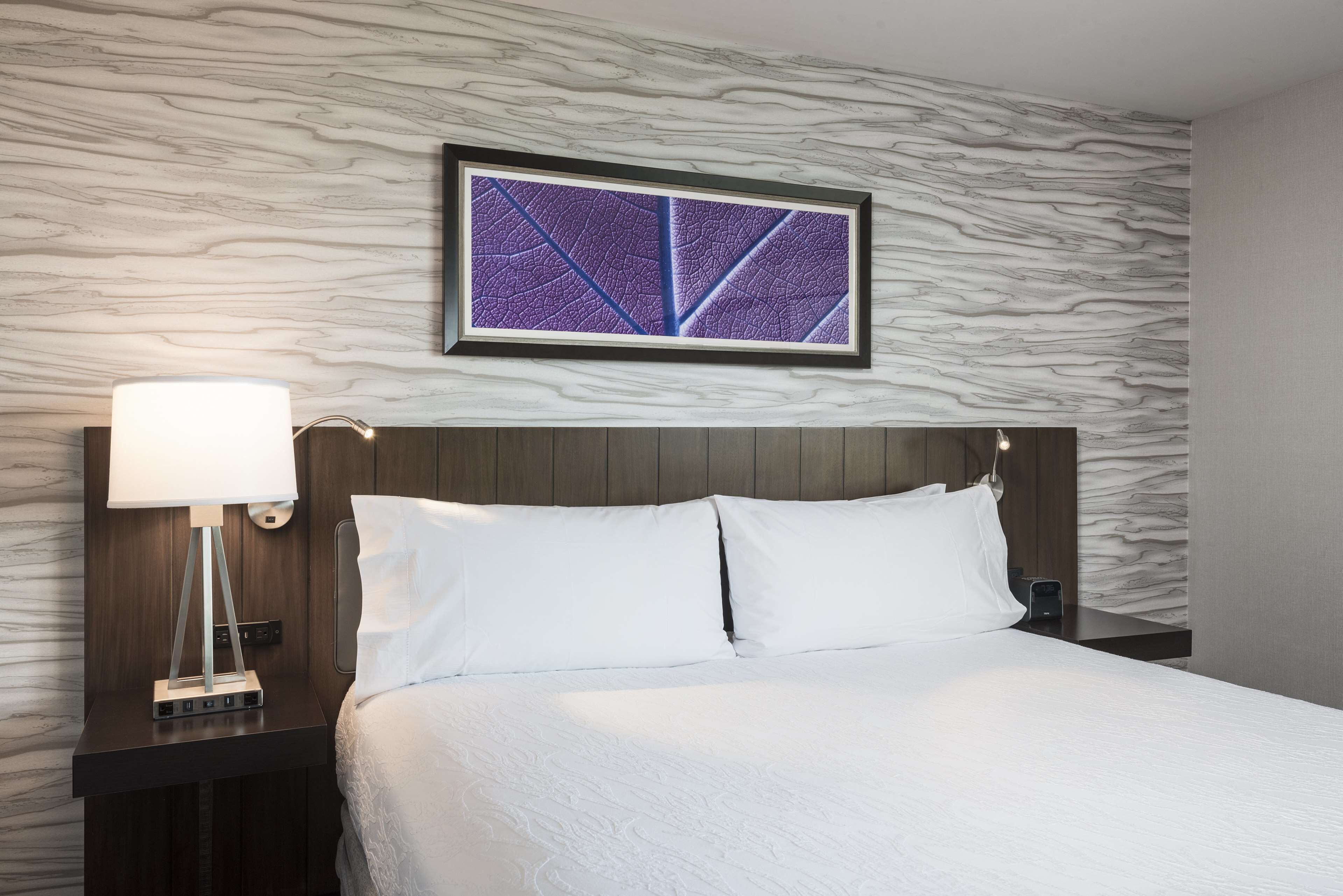 A neatly made bed with white linens, a modern bedside table with a lamp, and a wall-mounted artwork above the bed.