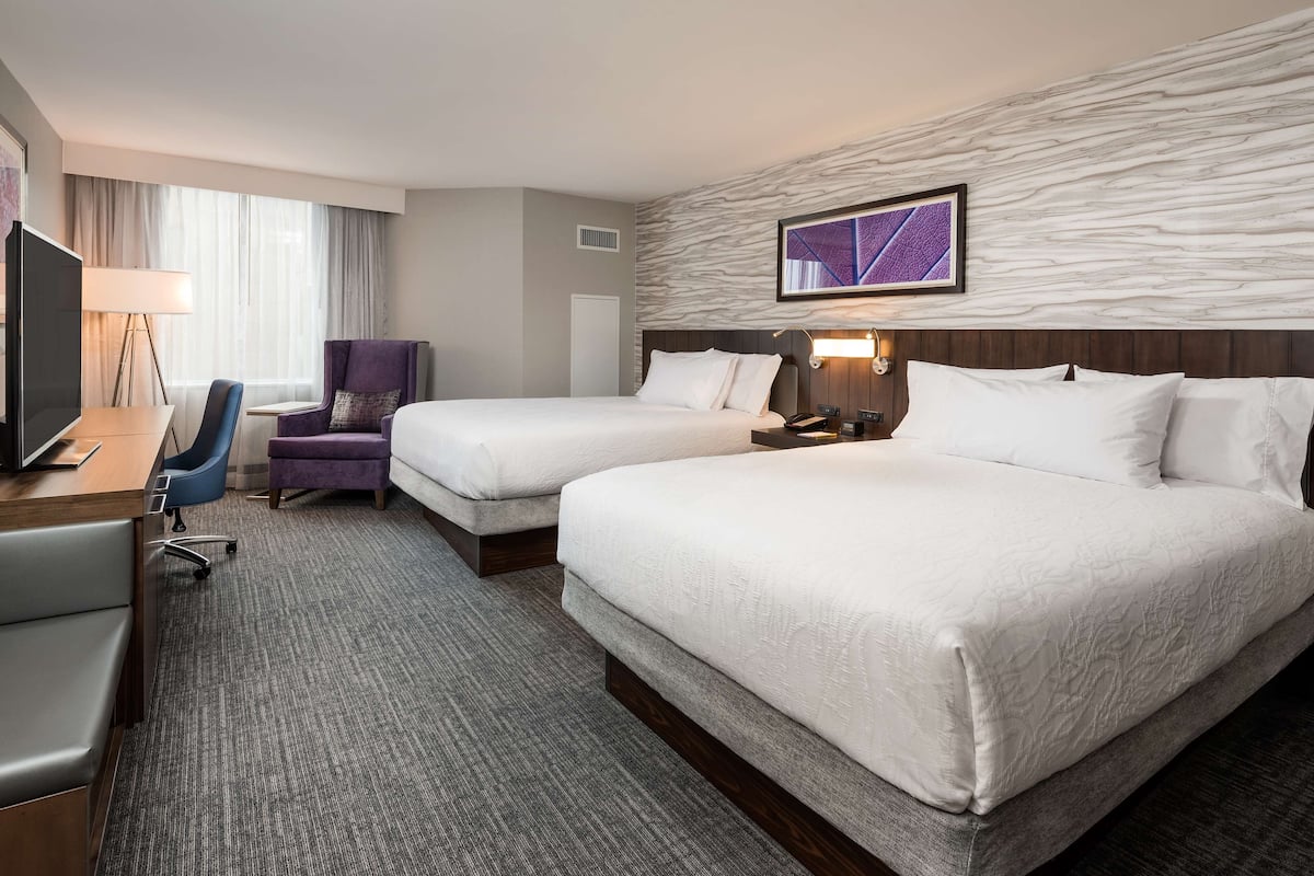 A hotel room with two beds, a desk with a TV, a chair, a lamp, and a wall-mounted artwork.