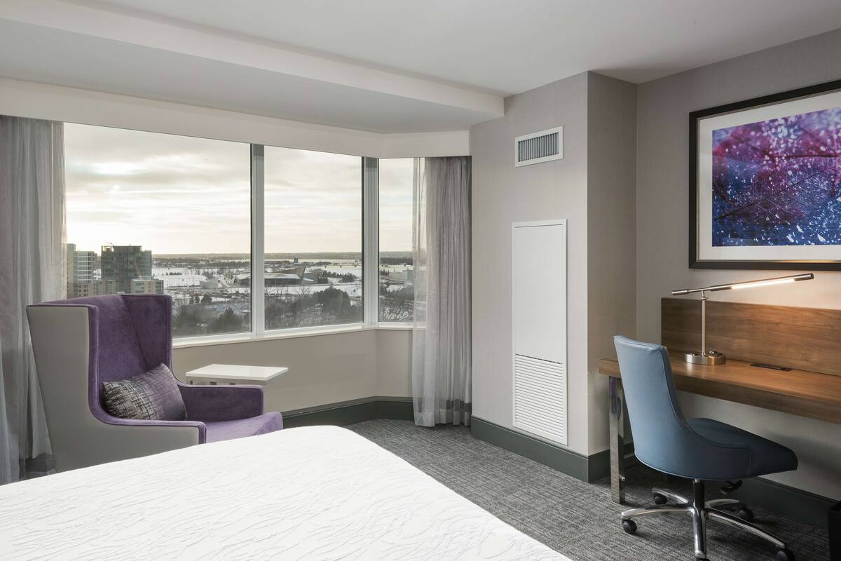 A hotel room with a large bed, a desk with a chair, a window with a view of the city, and a framed picture on the wall.
