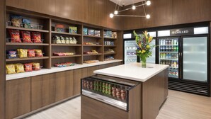 Un espace dédié aux collations et aux boissons, avec des étagères remplies de chips, un présentoir réfrigéré et un petit comptoir agrémenté d’un vase.