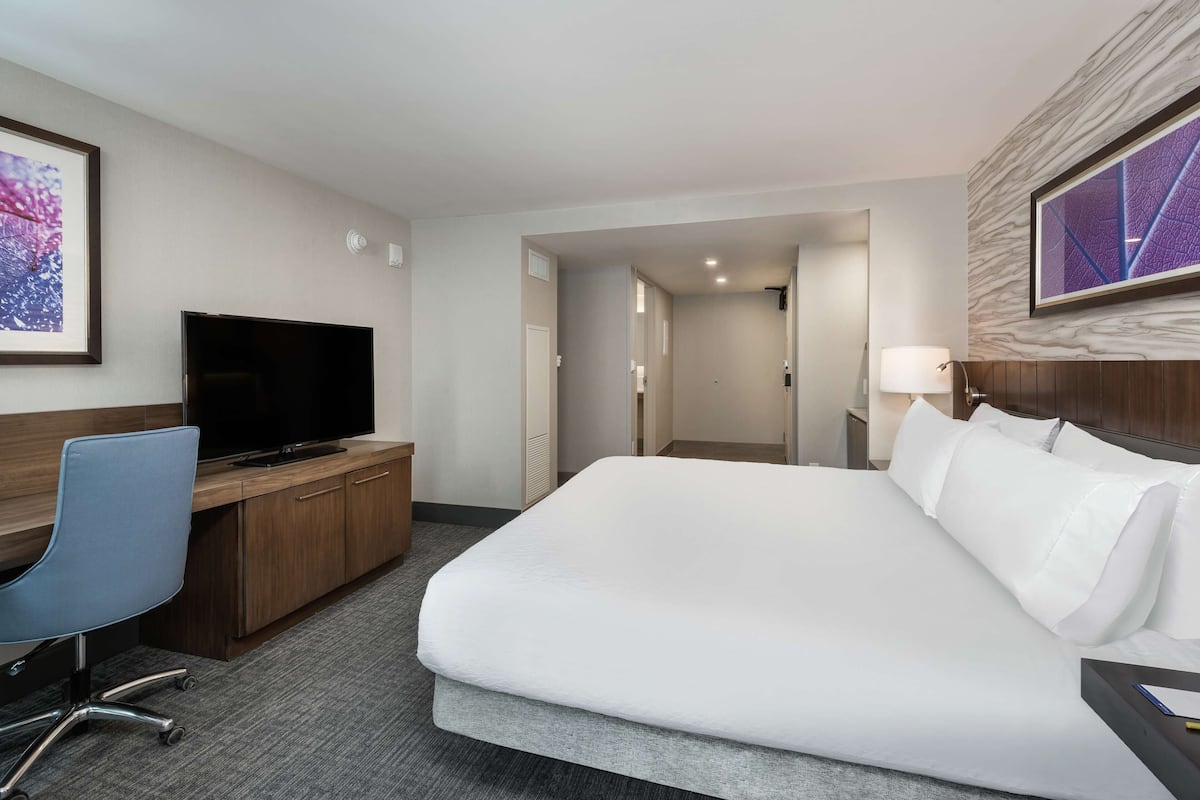 A hotel room with a large bed, a desk with a flat-screen TV, a chair, and a lamp.
