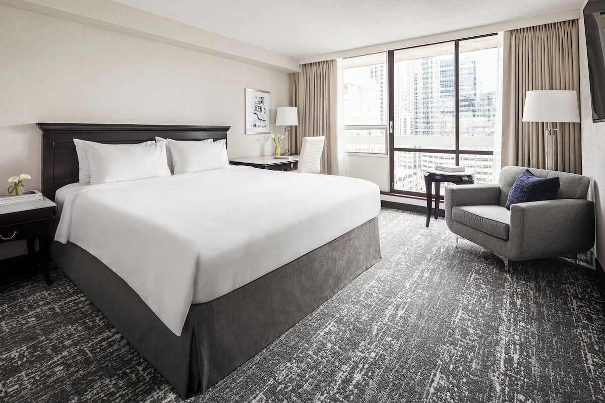 A hotel room with a large bed, a desk, a chair, and a view of the city through large windows.