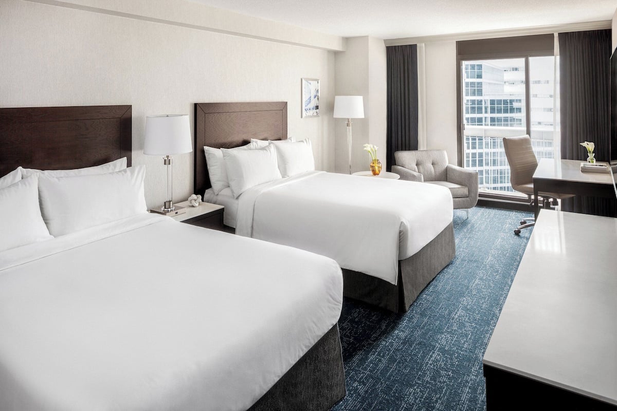 A hotel room with two beds, a desk, a chair, and a view of the city.
