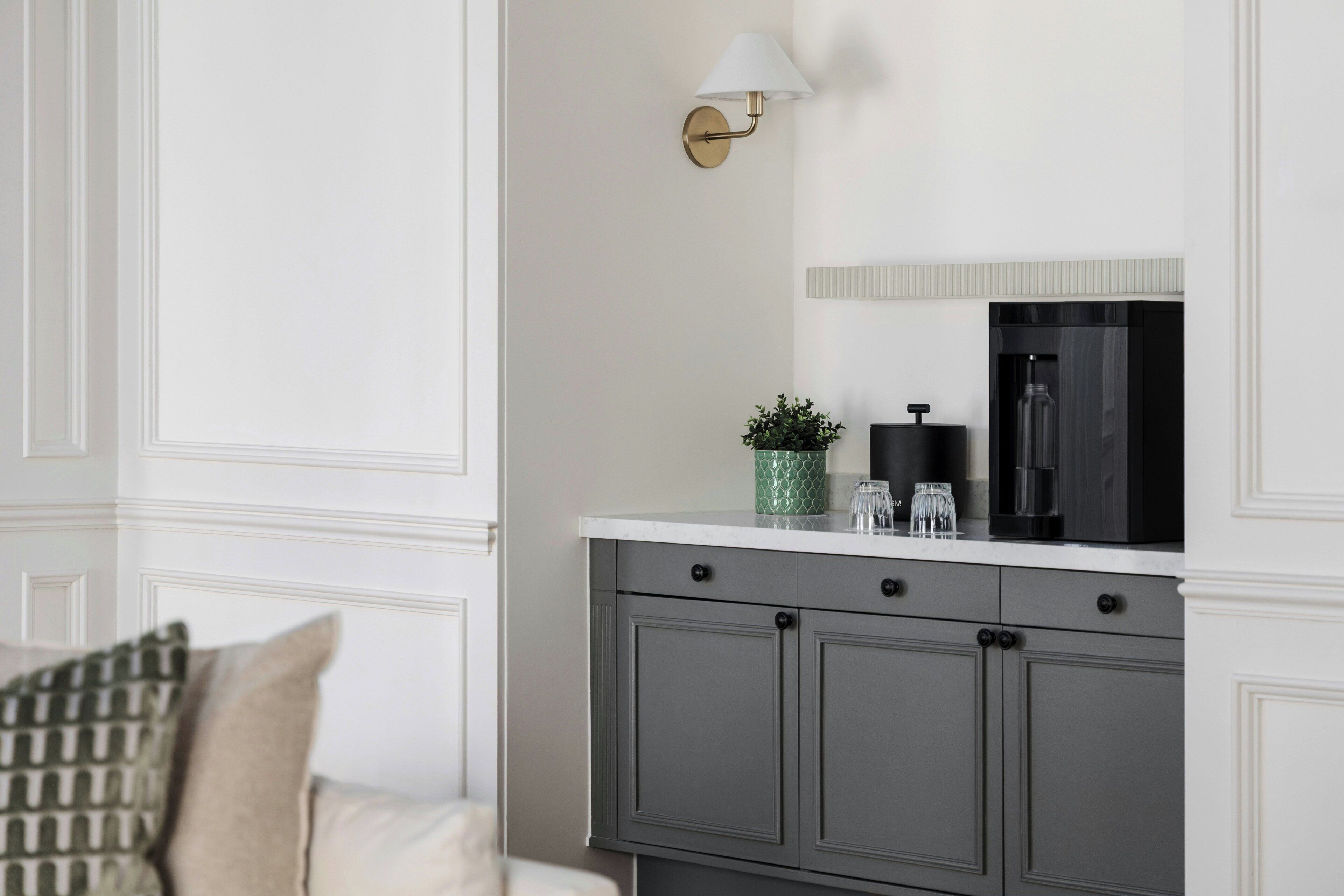 A modern kitchen with a white countertop, grey cabinets, a wall-mounted light fixture, and a small potted plant.