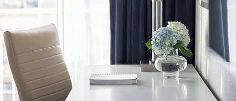 Habitación de hotel con escritorio, silla, lámpara, un jarrón con flores y ventana con cortinas.