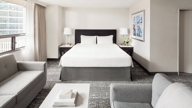 A hotel room with a large bed, two bedside tables with lamps, a sofa, a coffee table with books, and a window with curtains.