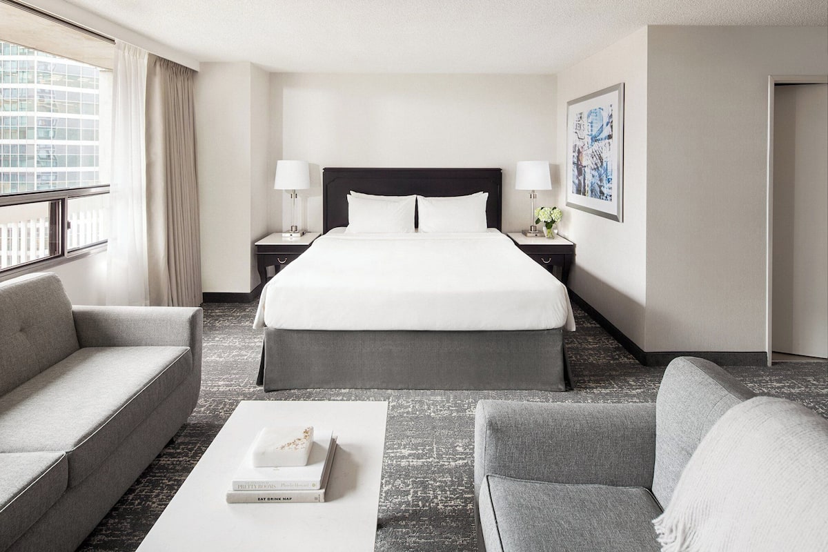 A hotel room with a large bed, two bedside tables with lamps, a sofa, a coffee table with books, and a window with curtains.