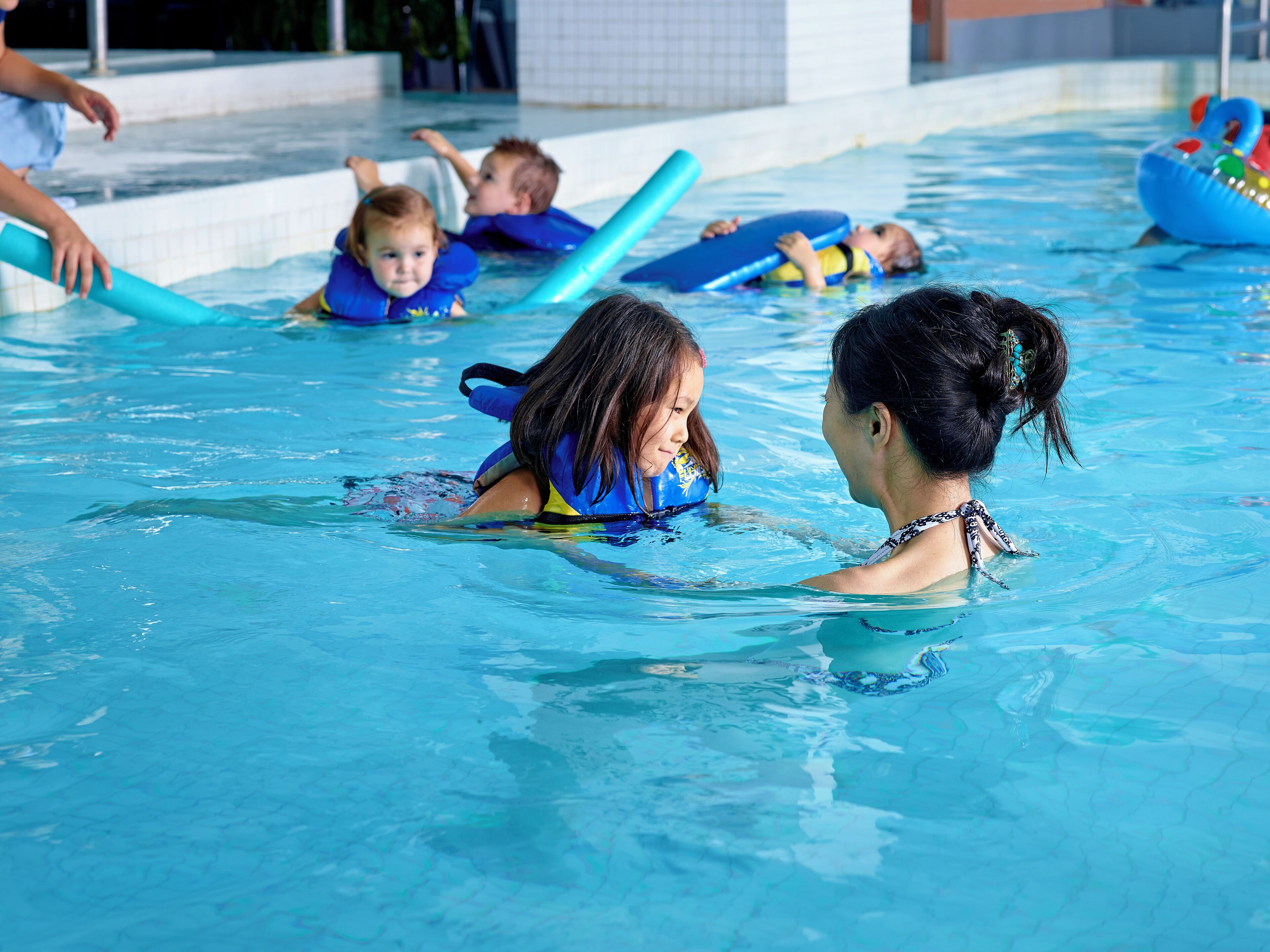 A swimming pool with children and adults in the water, some using floatation devices.
