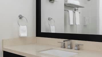 Un baño con un espejo grande, dos argollas para toallas, un lavamanos y una ducha.