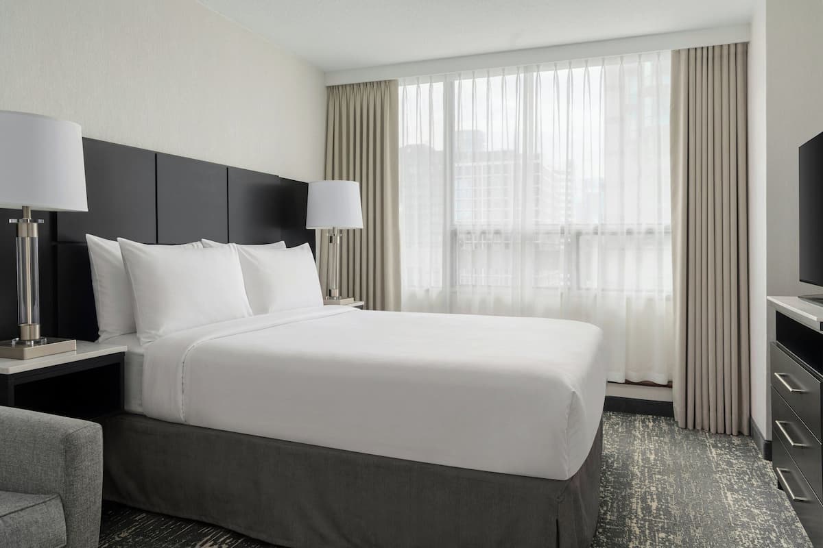 A hotel room with a large bed, two bedside tables, a TV, and a view of the city through the window.