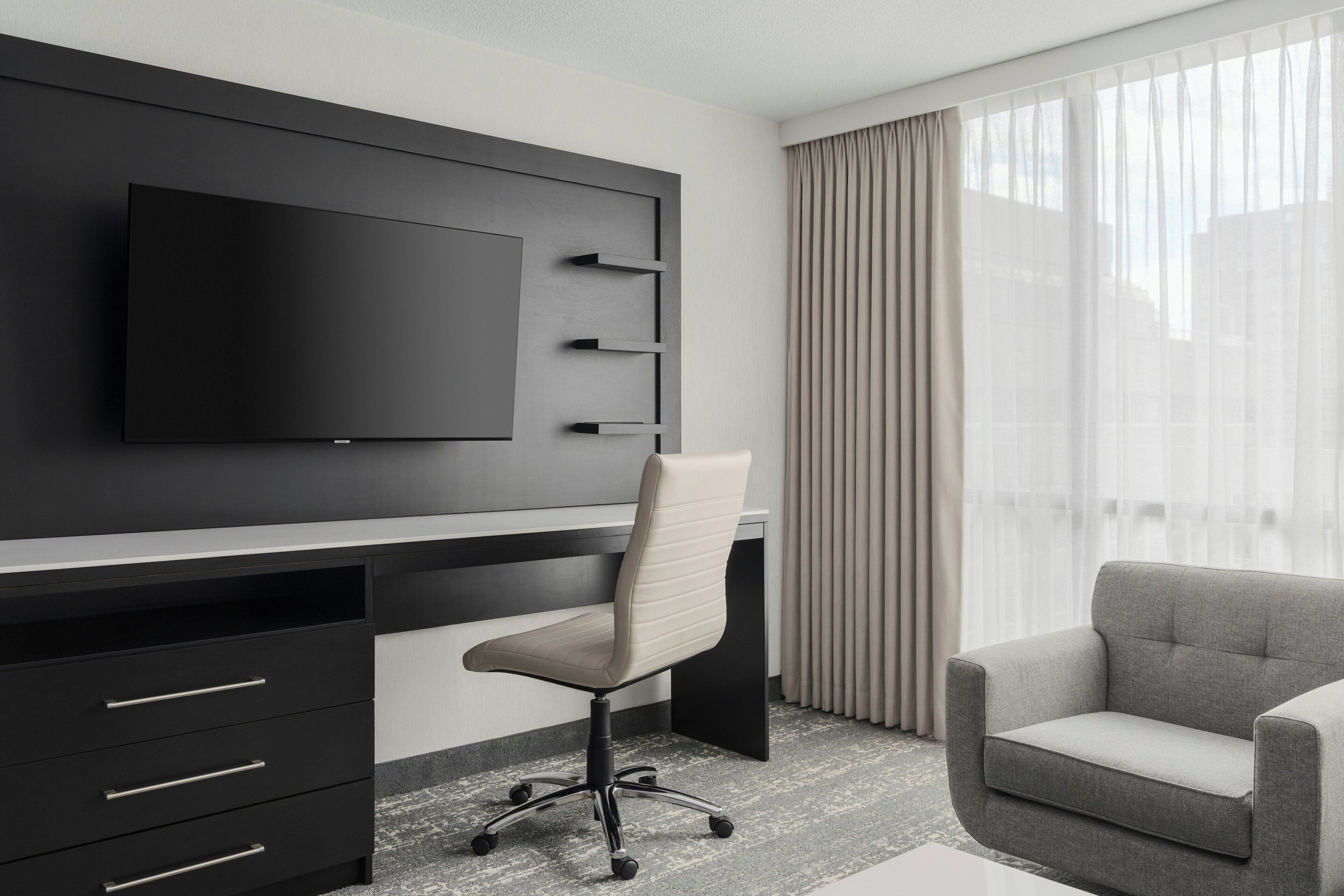 Una habitación de hotel moderna con una televisión de pantalla plana montada en la pared, un escritorio con cajones, una silla de oficina acolchada, un sofá y un amplio ventanal con cortinas translúcidas.