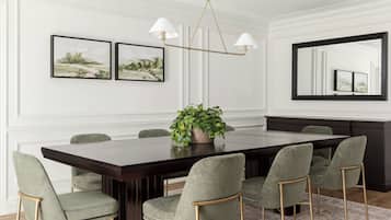A dining room with a long table, six chairs, a chandelier, and two framed paintings on the wall.