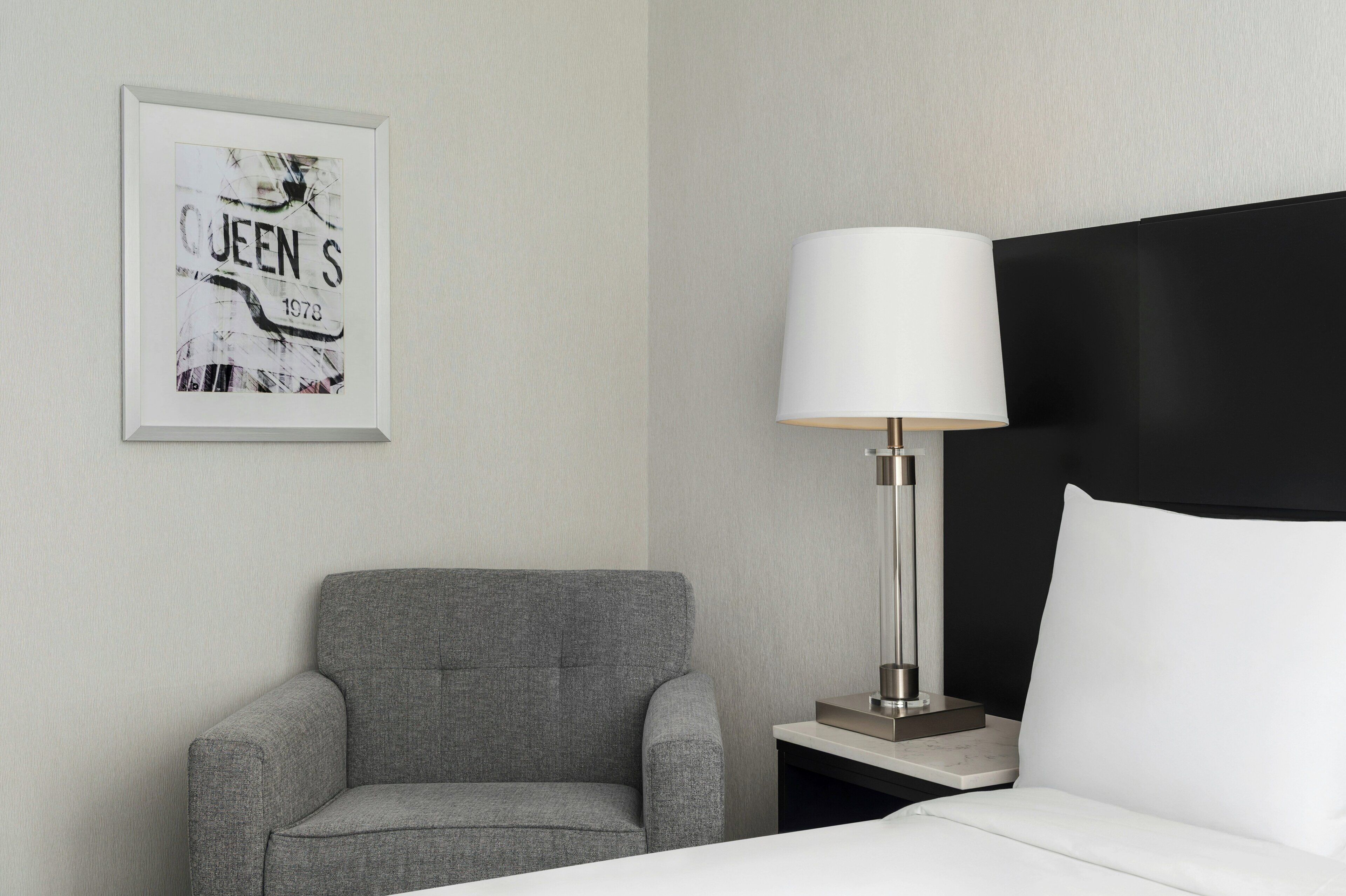 Habitación de hotel con un sillón gris, una mesita de noche con lámpara y un cuadro en la pared.