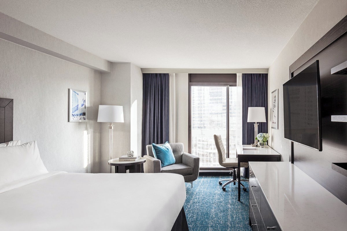 A hotel room with a large bed, a desk with a chair, a TV, and a view of the city through the window.
