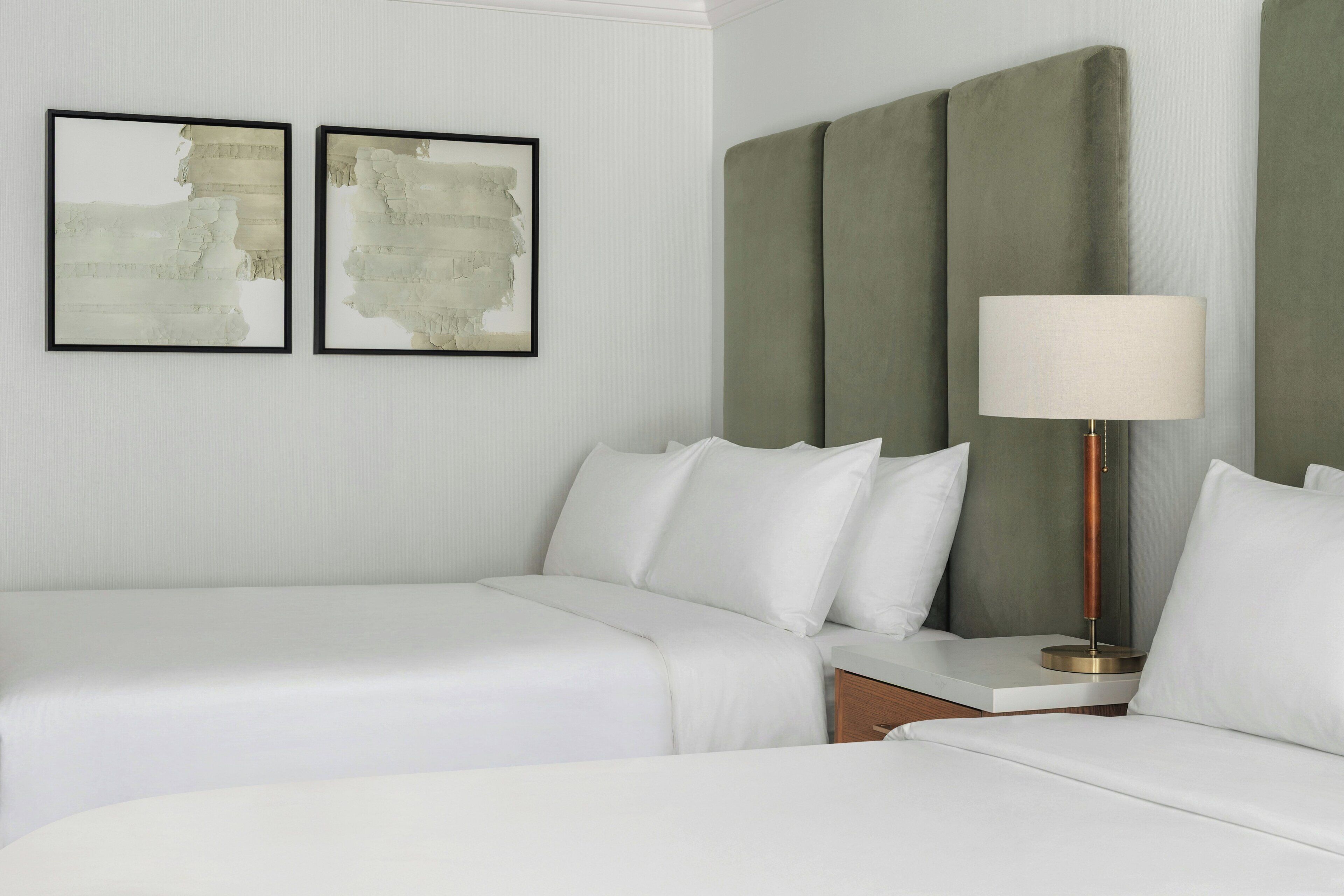 A hotel room with two beds, a nightstand, a lamp, and two abstract paintings on the wall.
