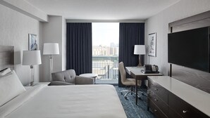 A hotel room with a large bed, a desk, a chair, a TV, and a view of the city through the window.