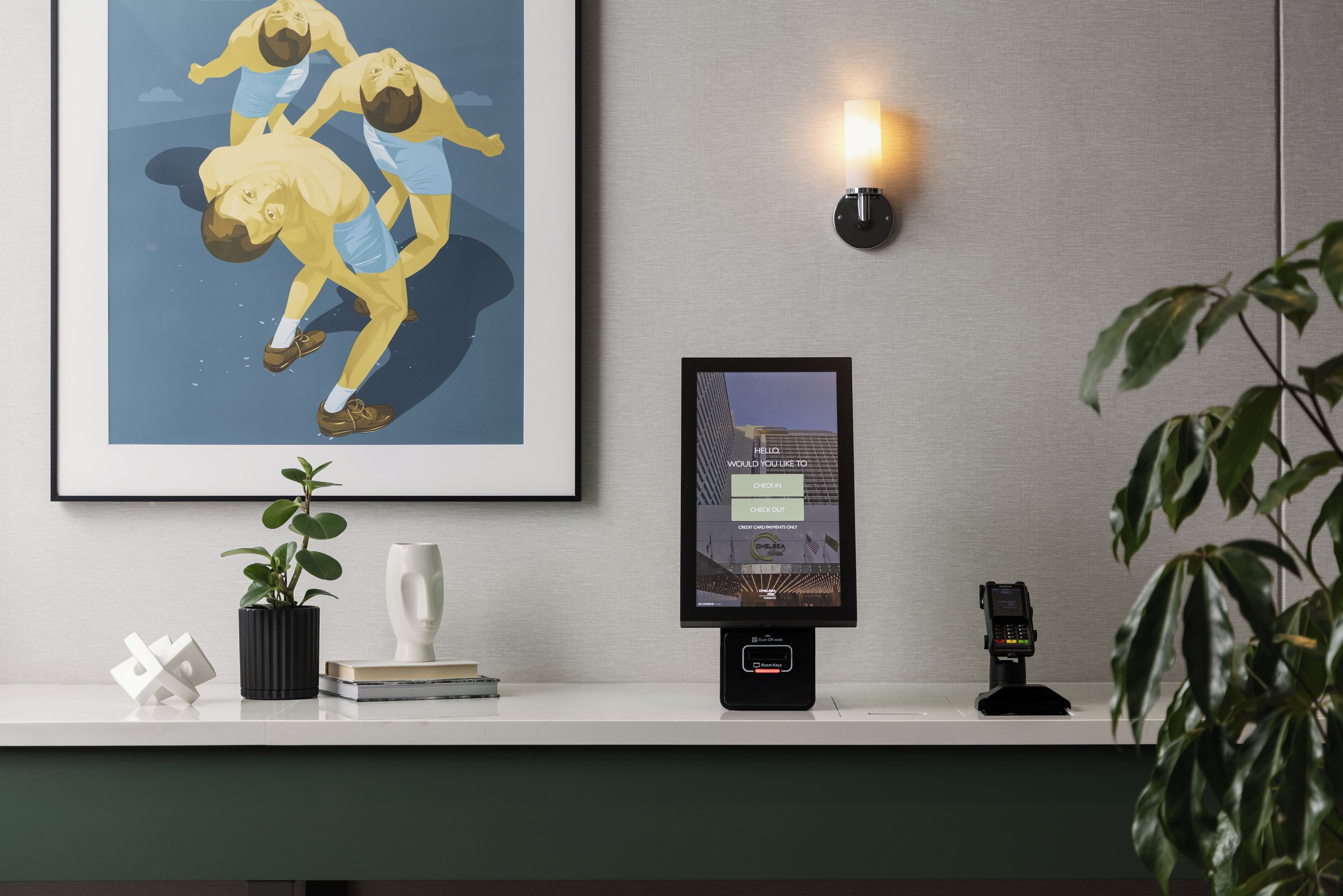A hotel reception desk with a framed artwork, a potted plant, a white vase, and a digital check-in kiosk.