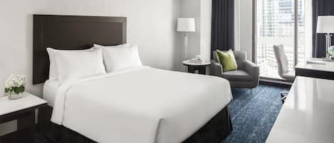 Habitación de hotel con una cama grande, un escritorio, una silla, una mesita de noche con una lámpara y vistas a la ciudad por la ventana.
