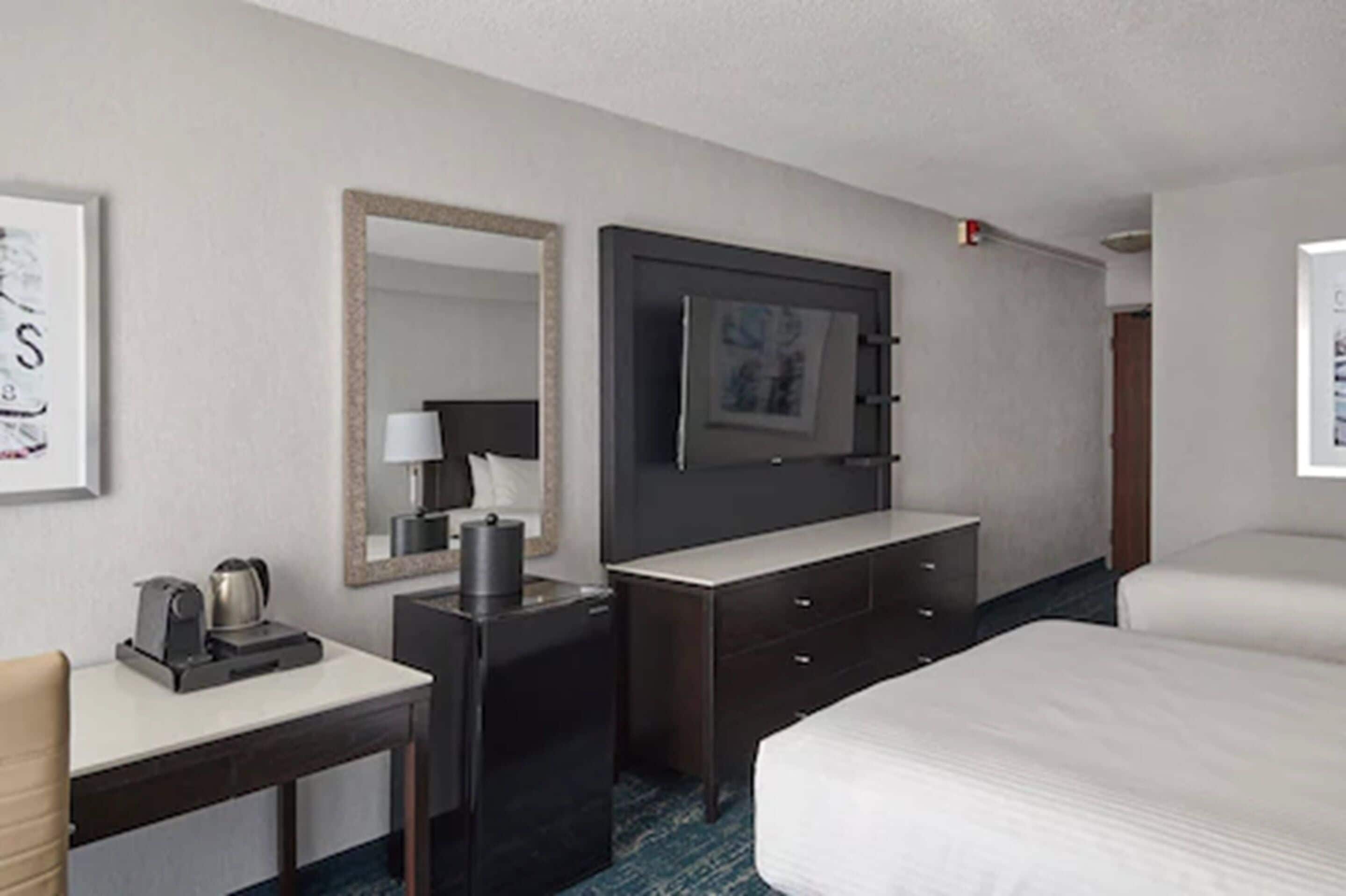 A hotel room with a bed, a desk with a coffee maker, a mirror, a television, and a dresser.