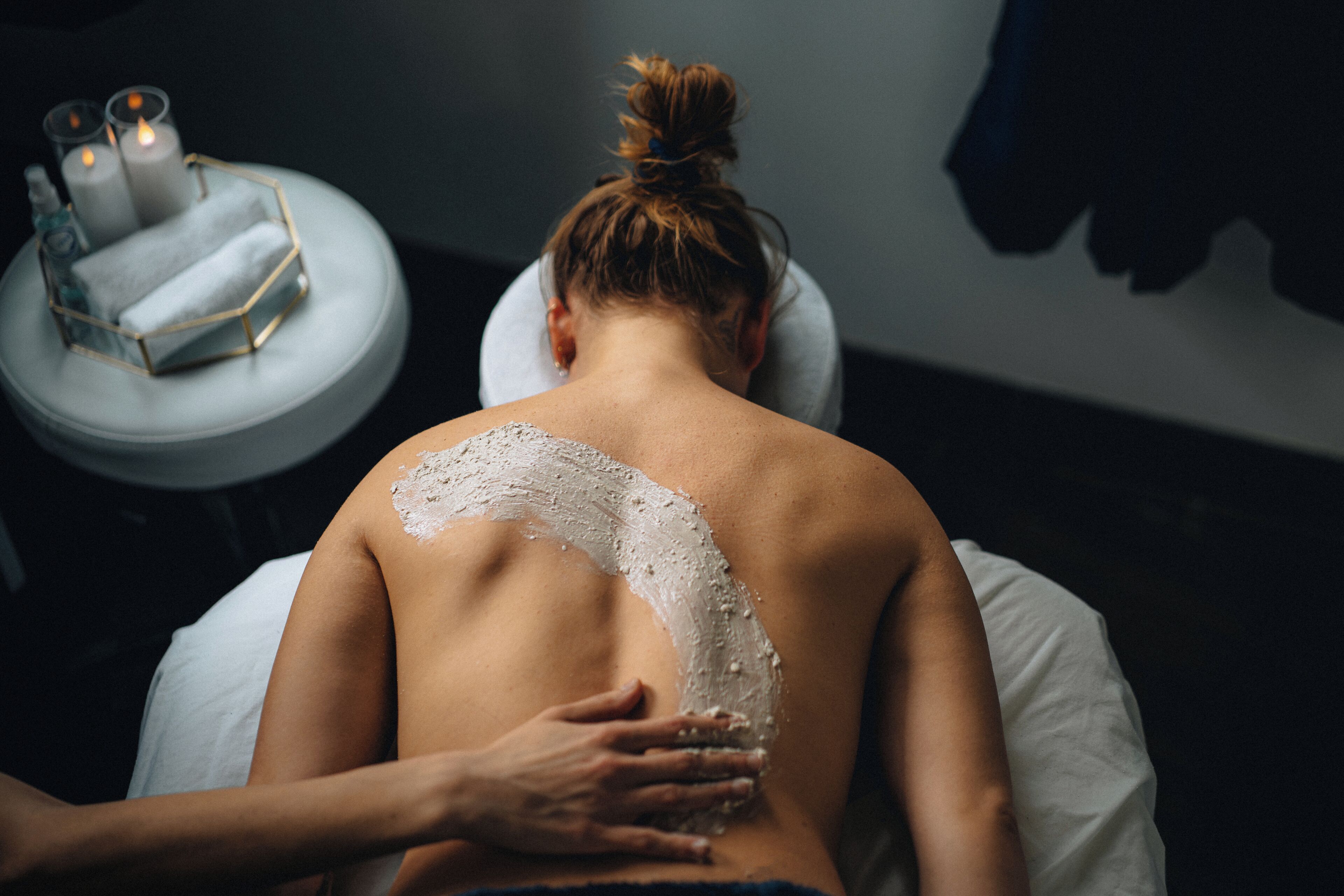 A person receiving a back treatment with a white substance, a candle, and a small table with a geometric container.