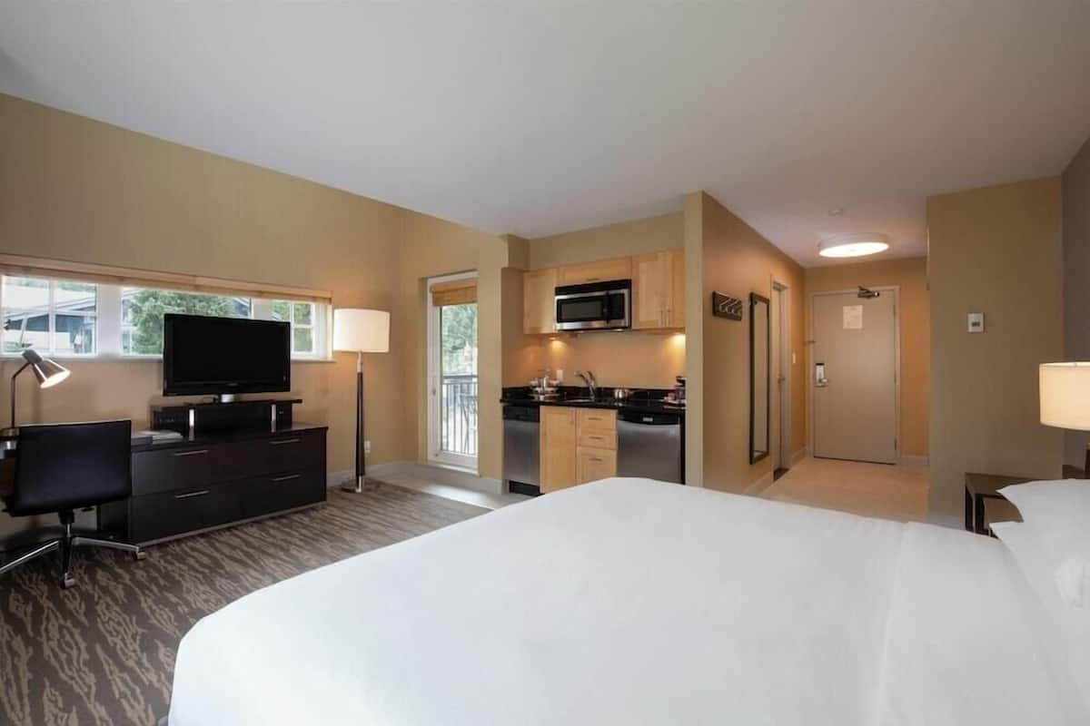 A hotel room with a bed, a television, a desk, a chair, a kitchenette, and a window.