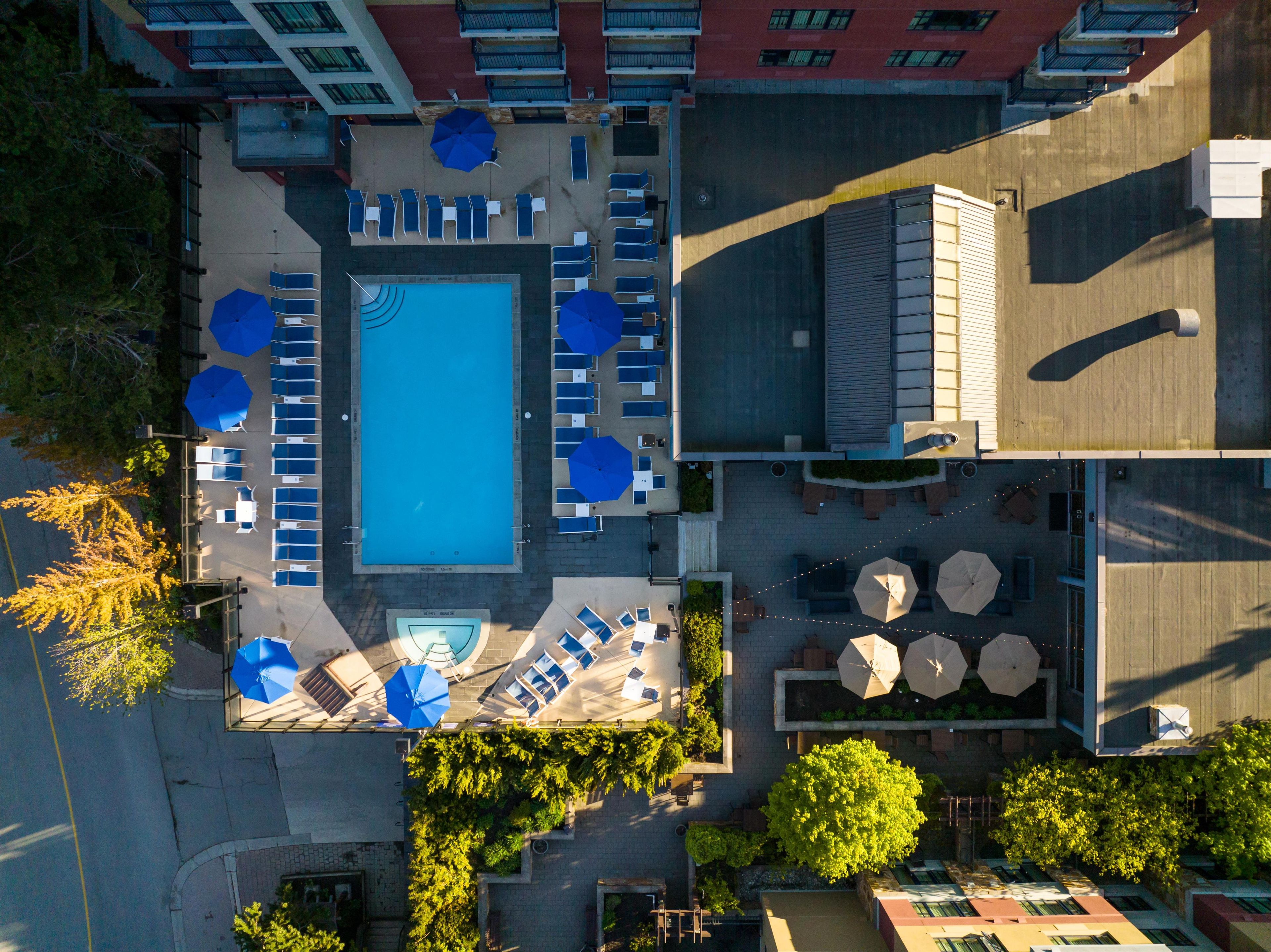 An aerial view of a hotel with a central swimming pool surrounded by lounge chairs and umbrellas, with trees and a building in the background.
