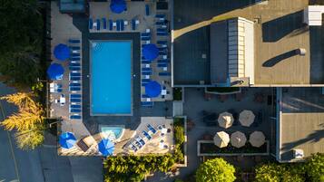 An aerial view of a hotel with a central swimming pool surrounded by lounge chairs and umbrellas, with trees and a building in the background.