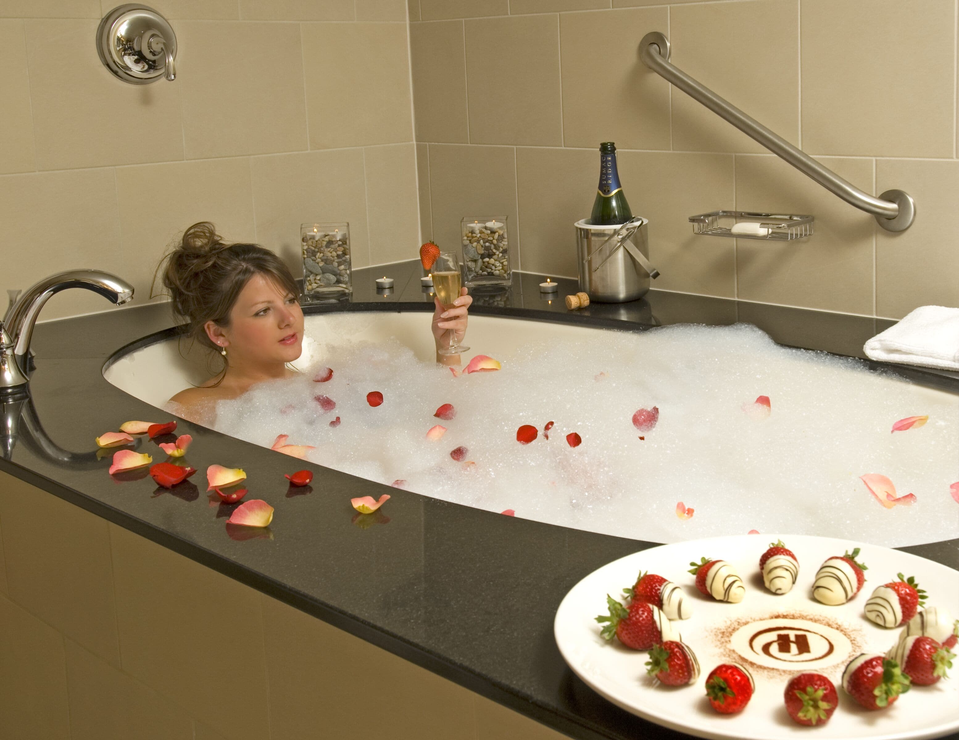 A bathtub filled with bubbles and rose petals, a bottle of champagne, and a plate of strawberries with chocolate.