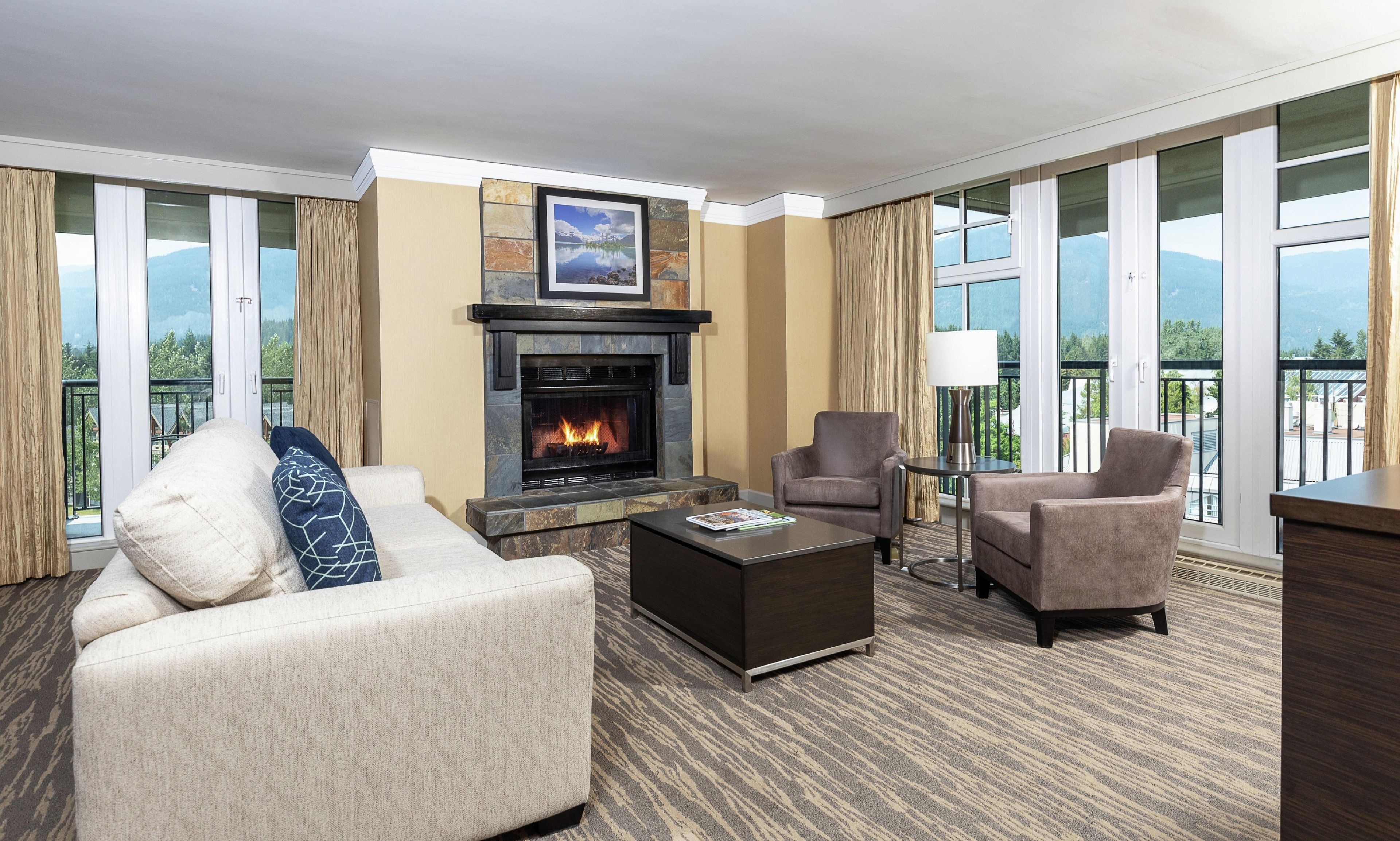 A hotel room with a fireplace, two armchairs, a sofa, a coffee table, and large windows with a mountain view.