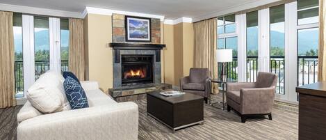 A hotel room with a fireplace, two armchairs, a sofa, a coffee table, and large windows with a mountain view.