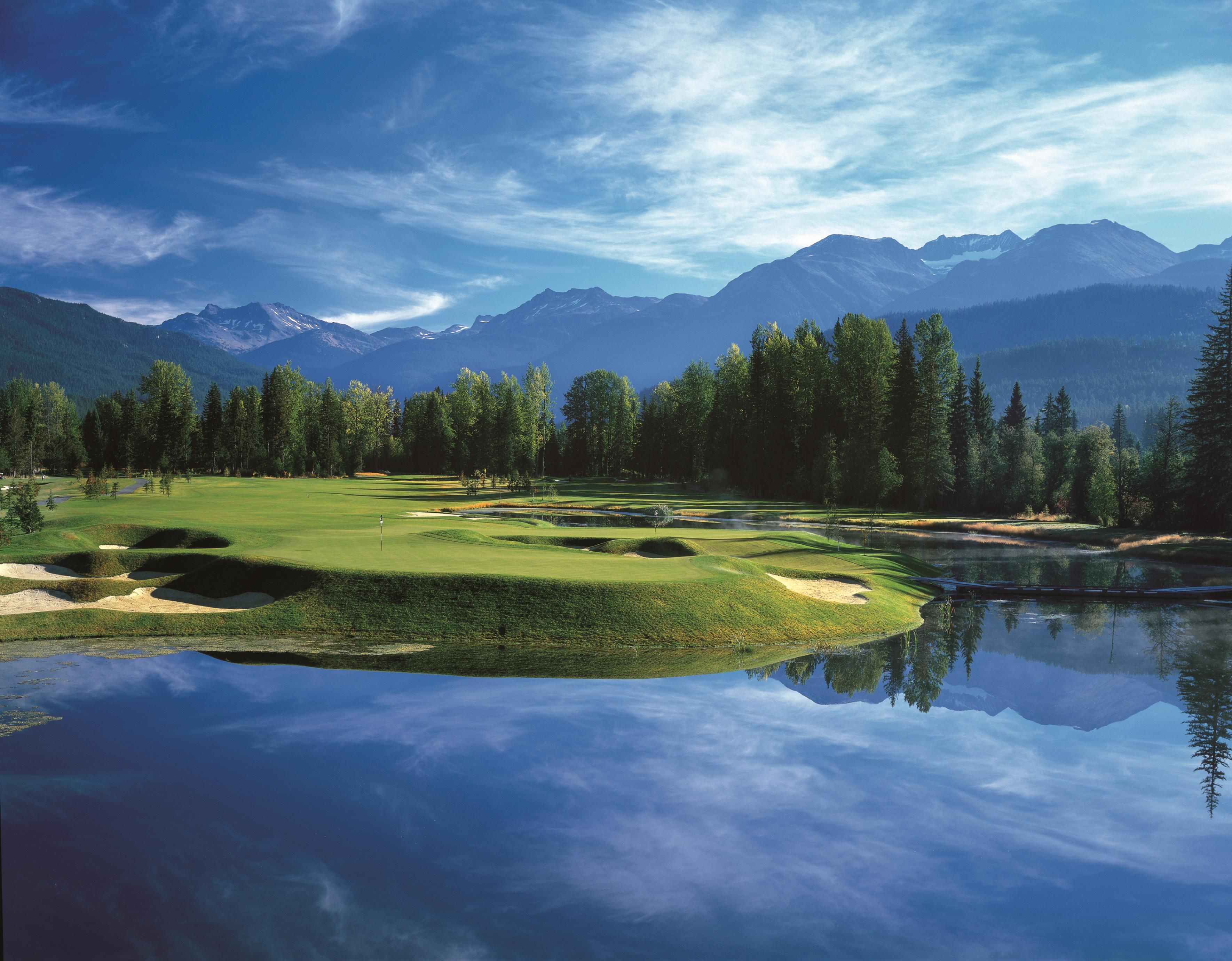 A serene golf course with a reflective pond, surrounded by dense forests and mountains in the background.