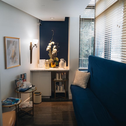 A compact living space with a blue sofa, a small table with a lamp, a framed picture, and a vase with flowers.