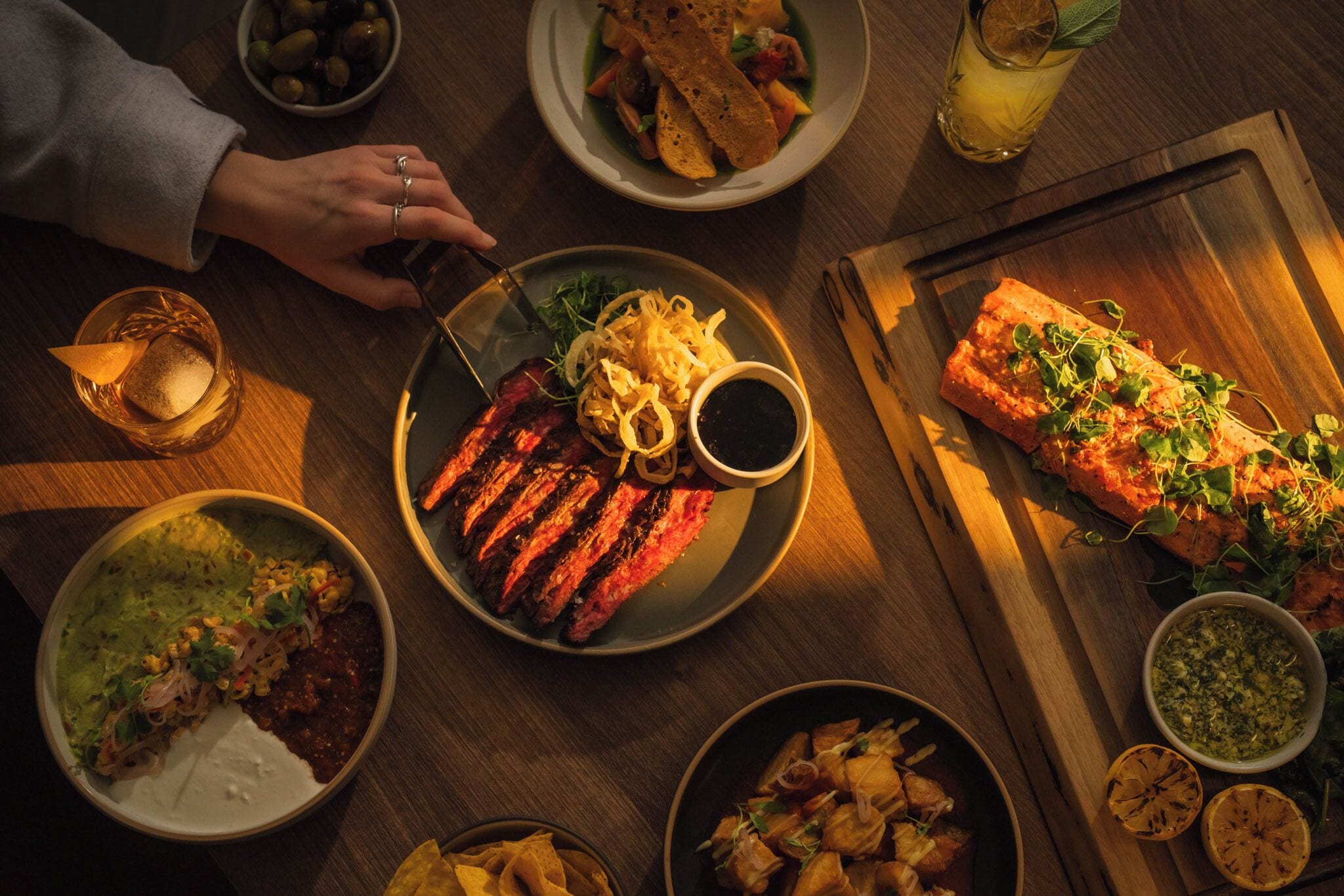 A dining table with various dishes, including grilled meat, salads, and a salmon fillet, along with drinks and a wooden cutting board.