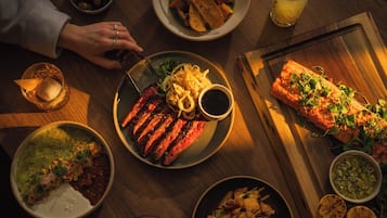 A dining table with various dishes, including grilled meat, salads, and a salmon fillet, along with drinks and a wooden cutting board.
