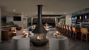 Un área lounge moderna con chimenea central, asientos cómodos y un bar con diversas bebidas.