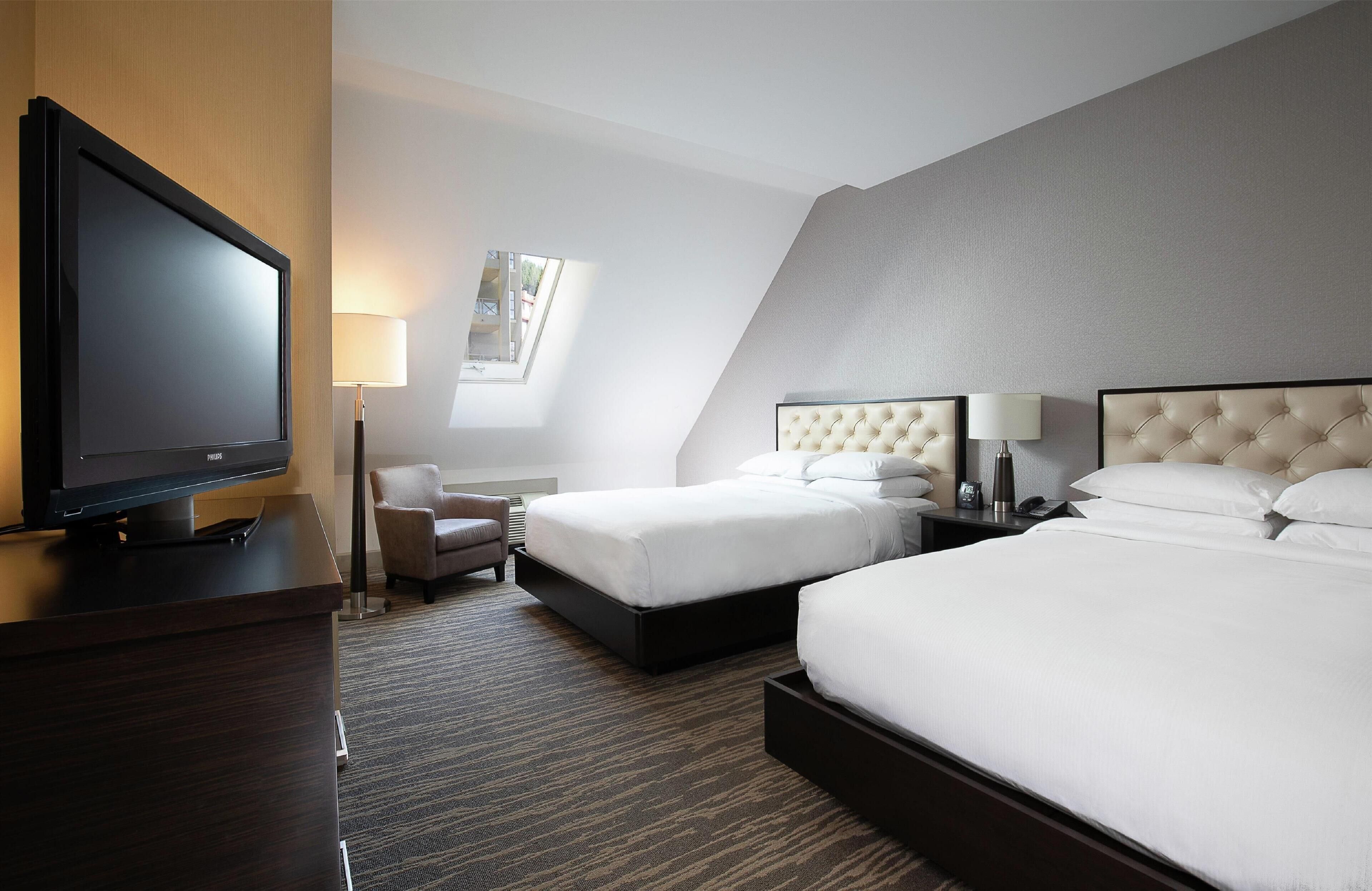 A hotel room with a flat-screen TV, two beds with tufted headboards, a desk, a chair, and a lamp.