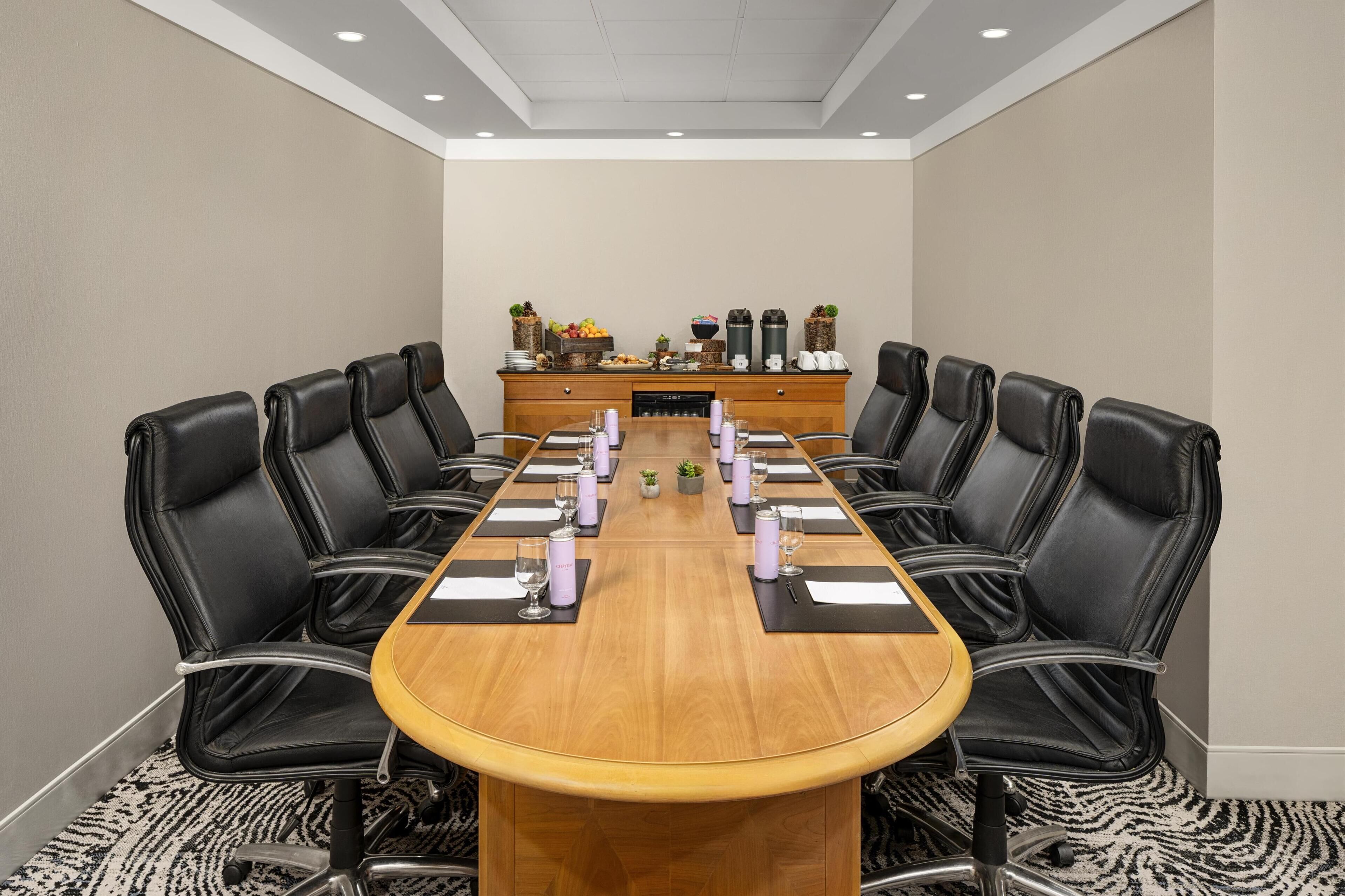 A conference room with a long wooden table, black leather chairs, and a buffet table with fruits and beverages.