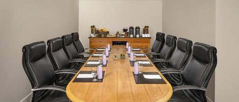 A conference room with a long wooden table, black leather chairs, and a buffet table with fruits and beverages.