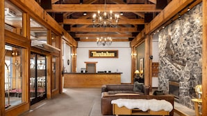A lobby with a reception desk, wooden beams, a stone wall, a fireplace, and a large chandelier. - Tantalus Resort Lodge (Whistler)