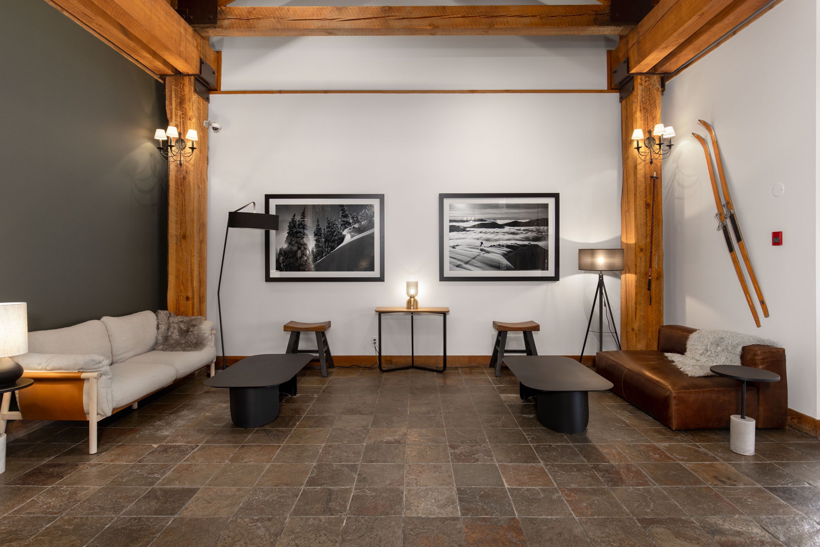Un salón moderno con suelo de baldosas de piedra, vigas de madera, dos fotografías enmarcadas, un sofá, una mesa de centro, una mesita con una lámpara y un par de esquíes apoyados en la pared.