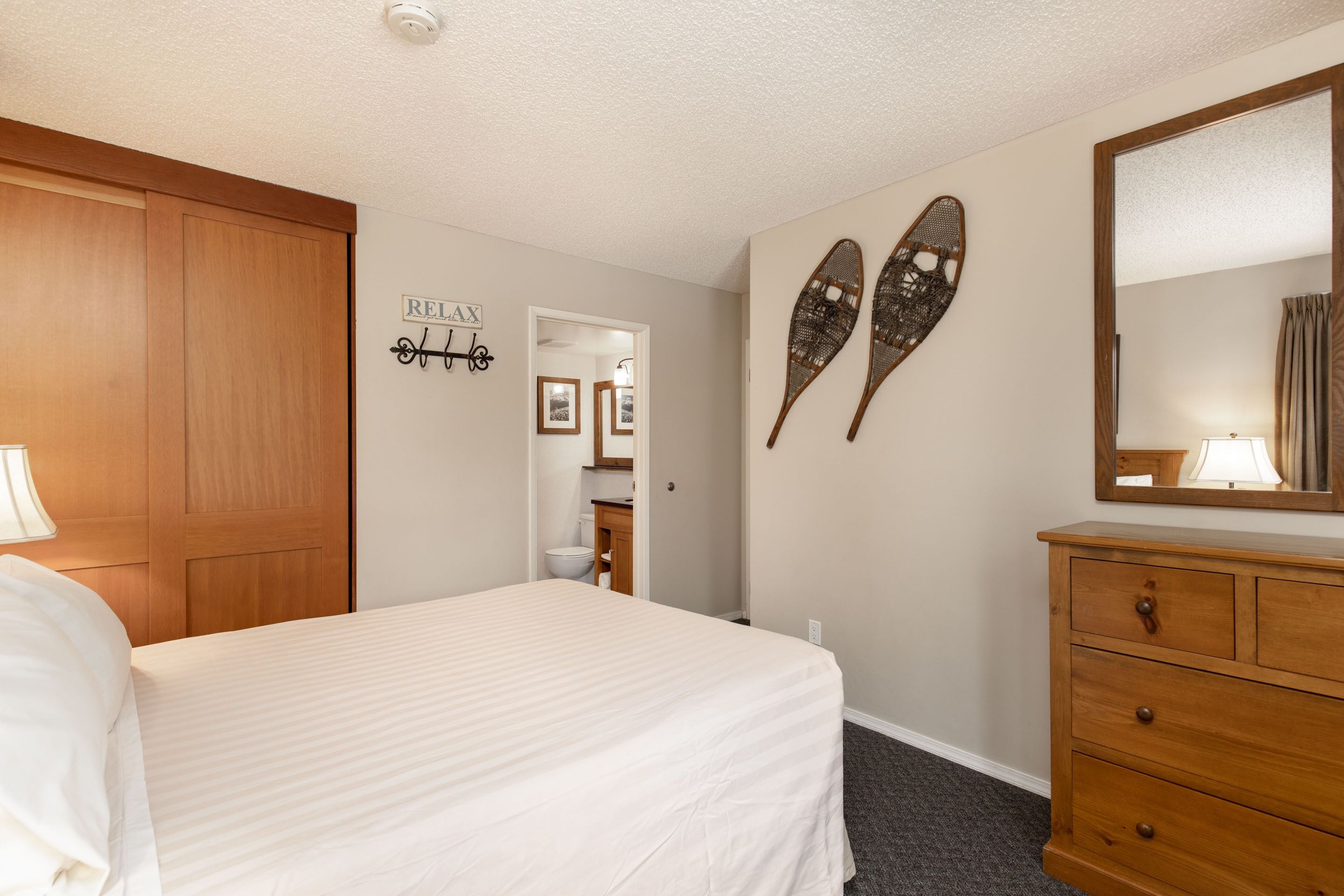 A bedroom with a bed, wooden wardrobe, a dresser, a mirror, and a wall decoration.