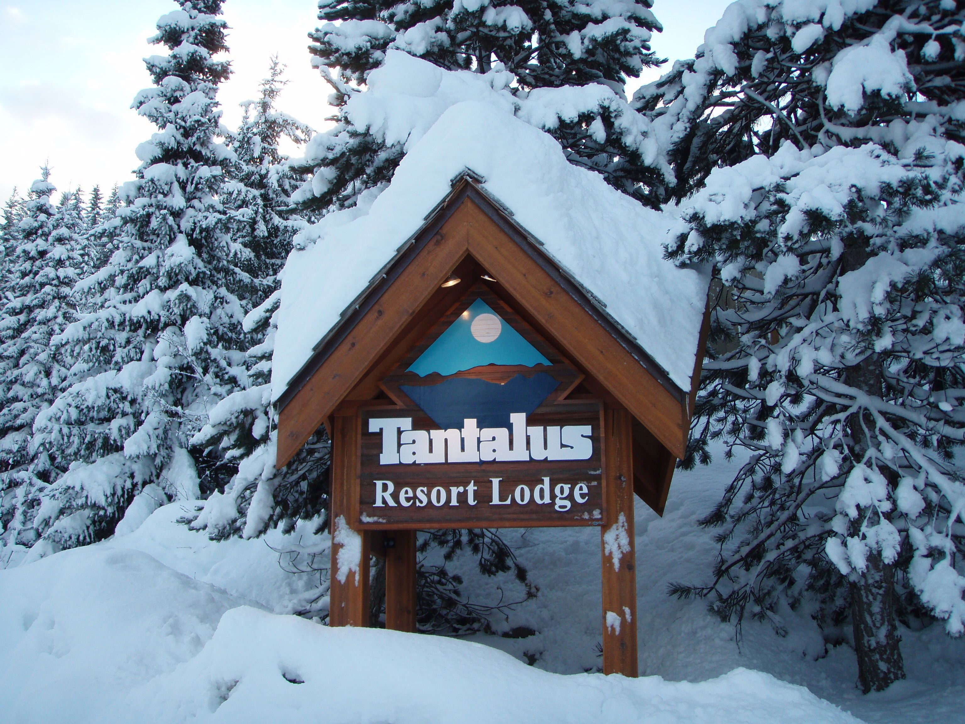 Lumihuntuun peittynyt metsä reunustaa Tantalus Resort Lodgen puista kylttiä.