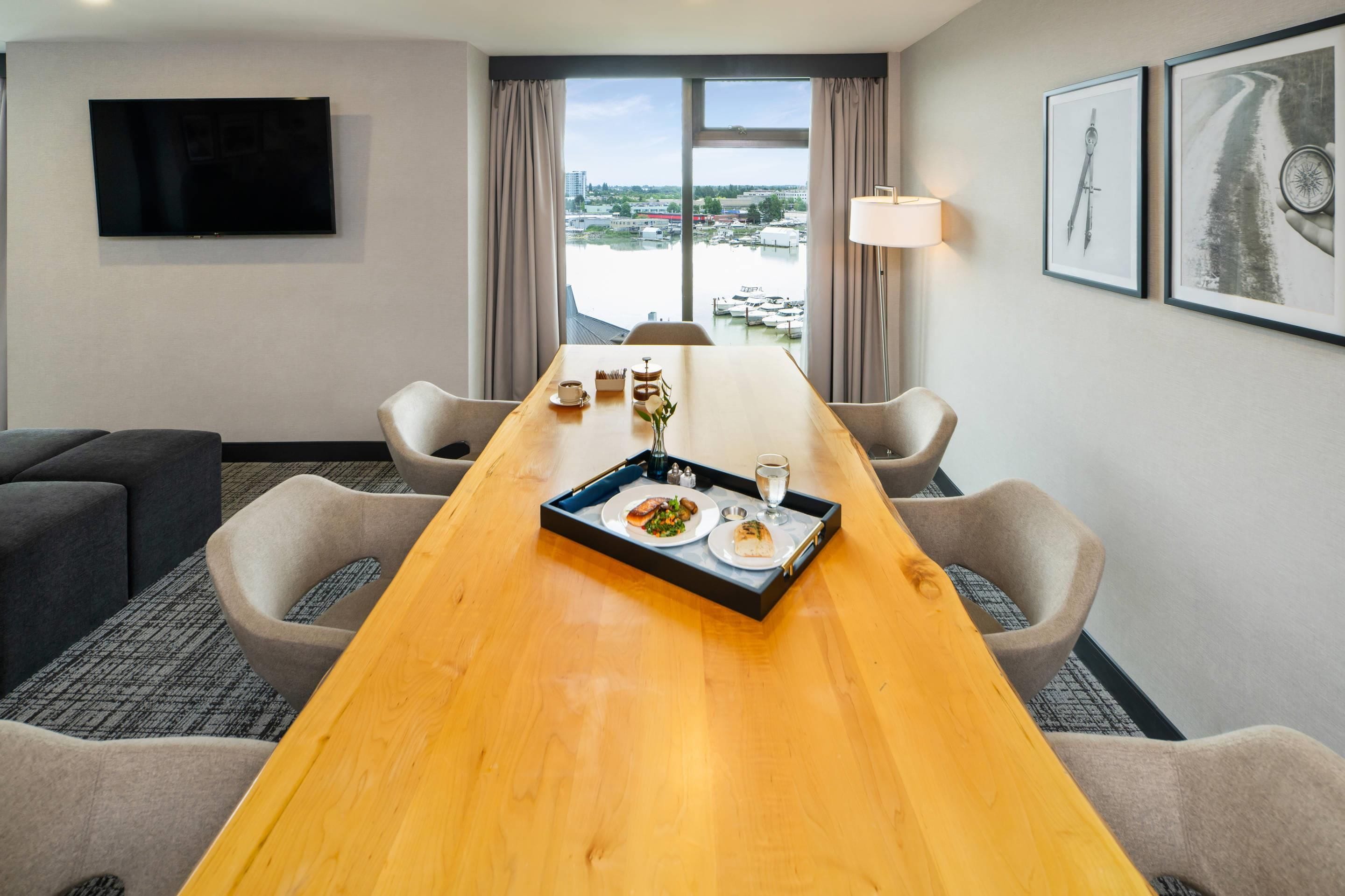 Una camera d'albergo con un tavolo da pranzo apparecchiato per due persone, una TV a schermo piatto montata a parete e vista sul porto turistico dalla finestra.